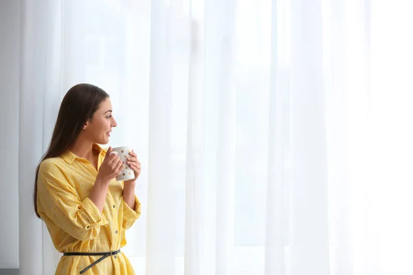 Young woman with cup near window indoors — 스톡 사진