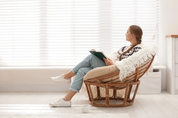 Evinin penceresinin yanındaki papasan sandalyesinde kitap okuyan genç bir kadın. — Stok fotoğraf