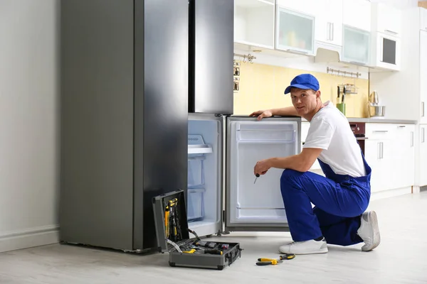 Male technician with screwdriver repairing refrigerator in kitch