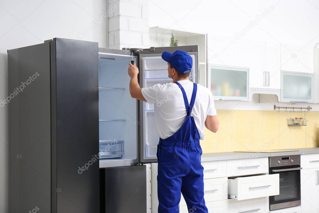 Male technician with screwdriver repairing refrigerator in kitch