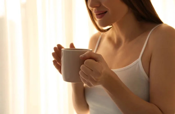 Young woman with cup near window at home, closeup. Lazy morning