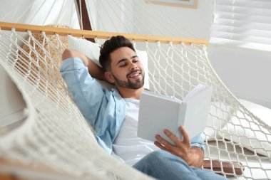 Genç adam evde hamakta kitap okuyor.