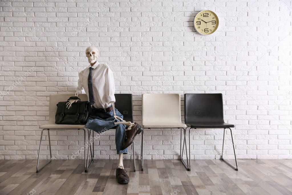 Human skeleton in office wear sitting on chair near brick wall indoors