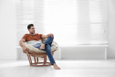 Attractive man relaxing in papasan chair near window at home. Sp clipart