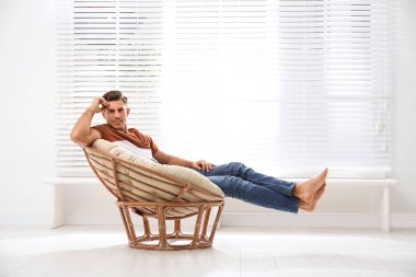 Attractive man relaxing in papasan chair near window at home clipart