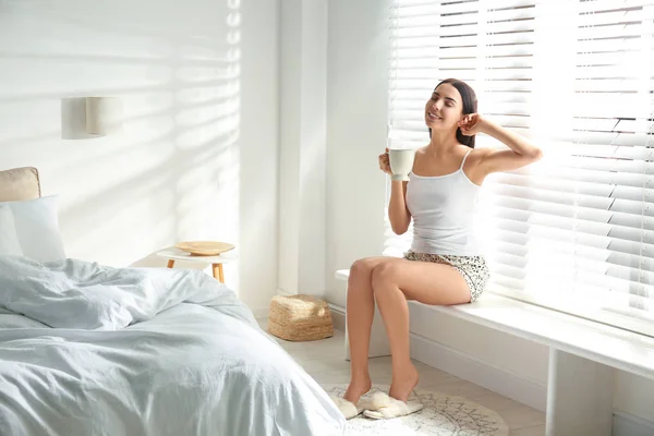 Νεαρή γυναίκα με φλιτζάνι τσάι κοντά στο παράθυρο. Τεμπέλικο πρωινό — Φωτογραφία Αρχείου