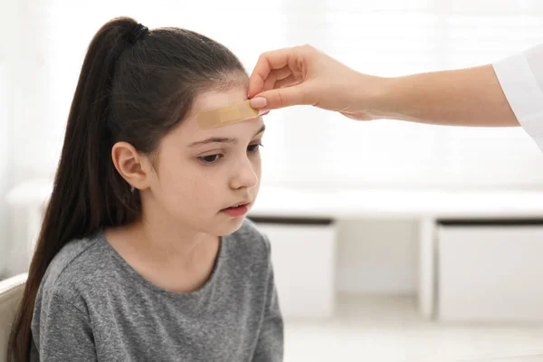 Doctor putting sticking plaster onto girl's forehead indoors — Stock Photo, Image