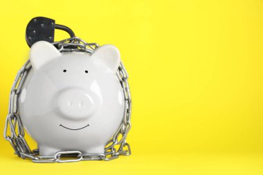 Piggy bank with steel chain and padlock on yellow background, space for text. Money safety concept