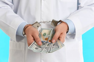 Açık mavi arka planda rüşvetle kelepçelenmiş bir doktor, yakın plan.