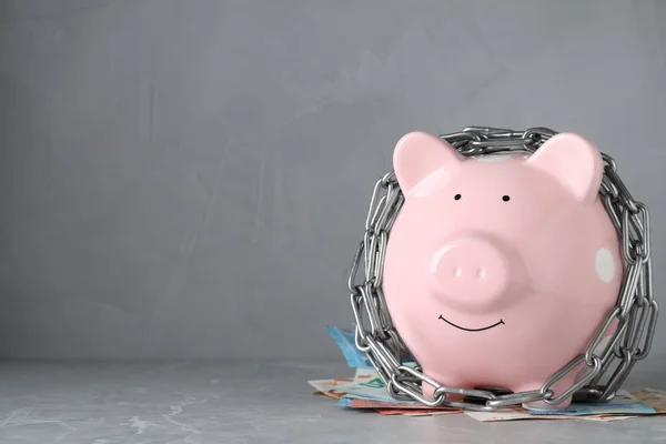 Piggy bank with steel chain and banknotes on grey marble table, space for text. Money safety concept