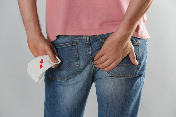 Man holding toilet paper with blood stain on light grey background, closeup. Hemorrhoid concept