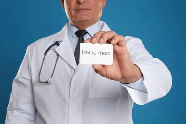 Doctor holding business card with word HEMORRHOID on blue background, closeup