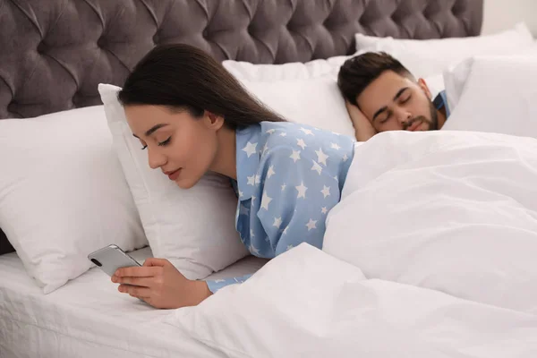 Jovem mulher usando smartphone enquanto seu namorado dorme na cama — Fotografia de Stock