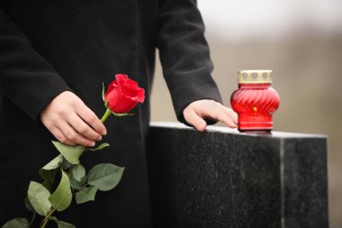 Woman holding red rose near black granite tombstone with candle 