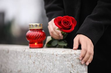 Woman holding red rose near grey granite tombstone with candle o