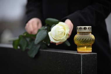 Woman holding white rose near black granite tombstone with candl