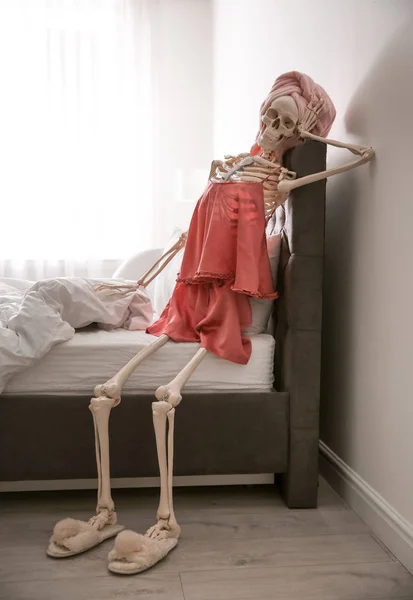 Human skeleton in silk pajamas and towel sitting on bed indoors