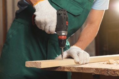 Professional carpenter twisting screw into wooden plank with drill in workshop, closeup