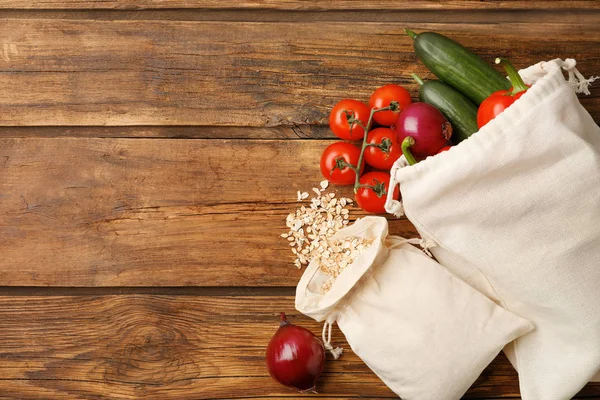 Cotton eco bags with vegetables and oat flakes on wooden table, flat lay. Space for text