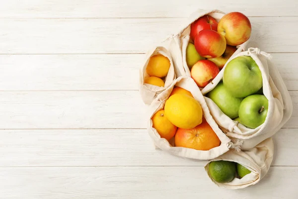 Cotton eco bags with fruits on white wooden table, flat lay. Space for text