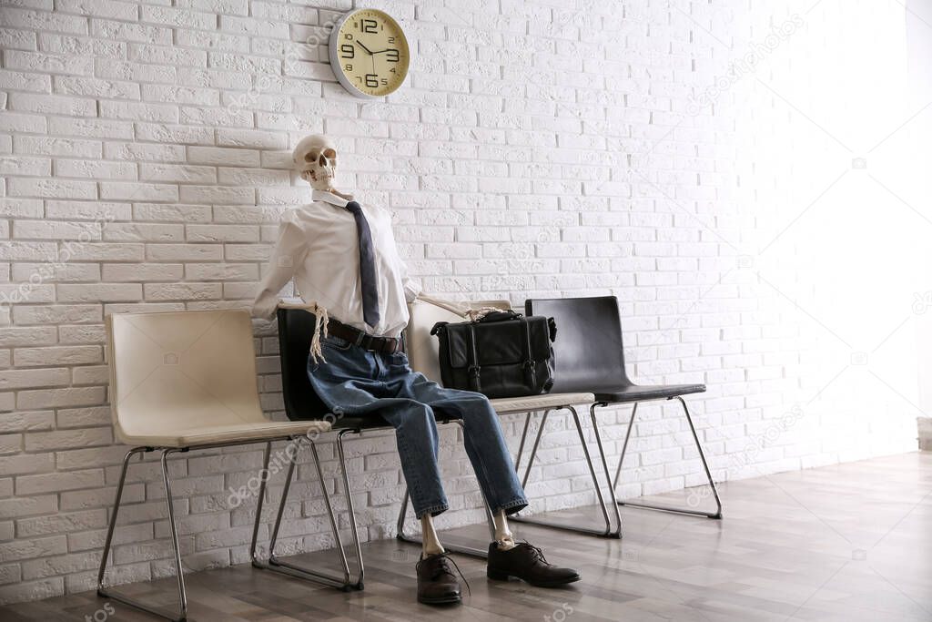 Human skeleton in office wear sitting on chair near brick wall indoors