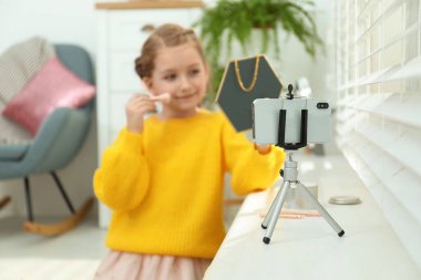 Cute little blogger with cosmetics recording video at home, focus on phone