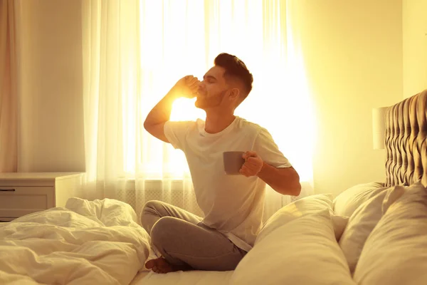 Young man with cup of coffee on bed at home. Lazy morning