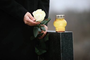 Woman holding white rose near black granite tombstone with candl
