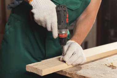 Professional carpenter twisting screw into wooden plank with drill in workshop, closeup