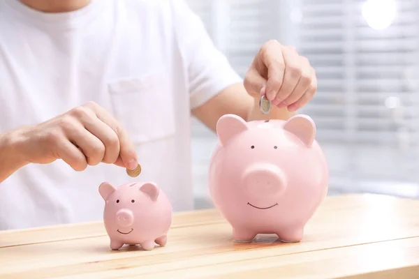 Man putting coins into piggy banks at wooden table, closeup — Stock Photo, Image