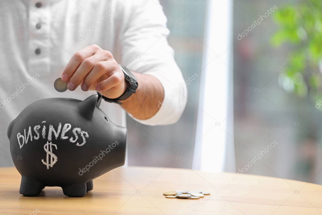 Man putting money into piggy bank with word BUSINESS at table, closeup. Space for text