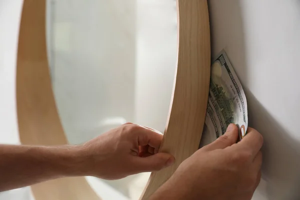 Man hiding money behind mirror indoors, closeup. Financial savin