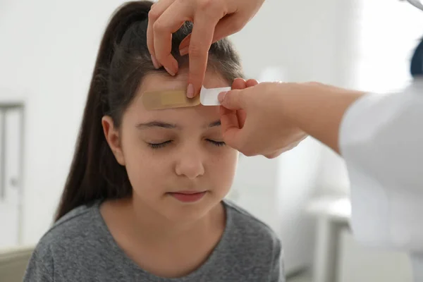 Doctor putting sticking plaster onto girl\'s forehead indoors