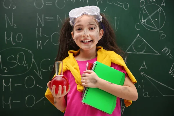 Schoolgirl holding flask and notebooks near chalkboard with chem — Stock Photo, Image