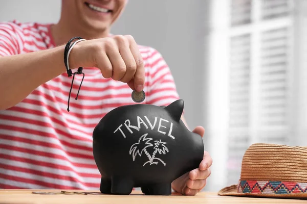Man putting coin into piggy bank with word TRAVEL at table, clos — Stock Photo, Image