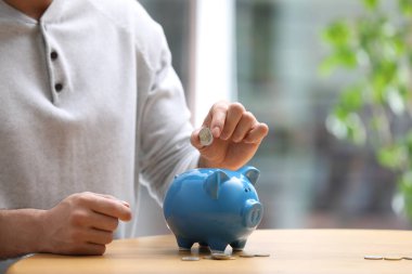 Man putting money into piggy bank at table, closeup. Space for text clipart