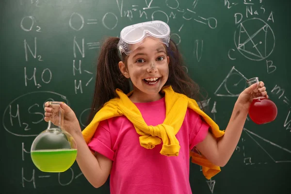 Schoolgirl holding flasks near chalkboard with chemical formulas — Stock Photo, Image
