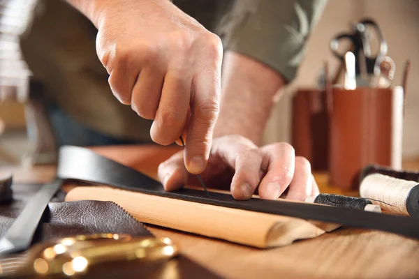 Man making holes in leather belt with stitching awl at table, closeup