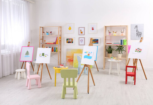 Easels with paintings and chairs for children in room