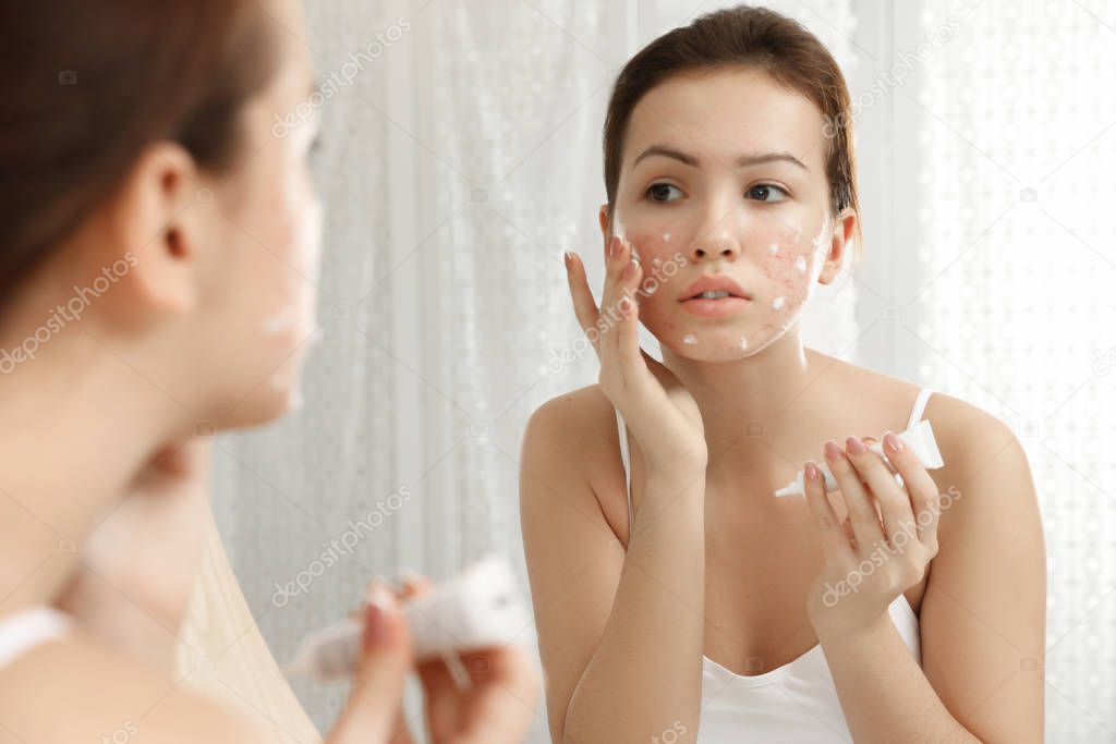 Teen girl with acne problem applying cream near mirror in bathro
