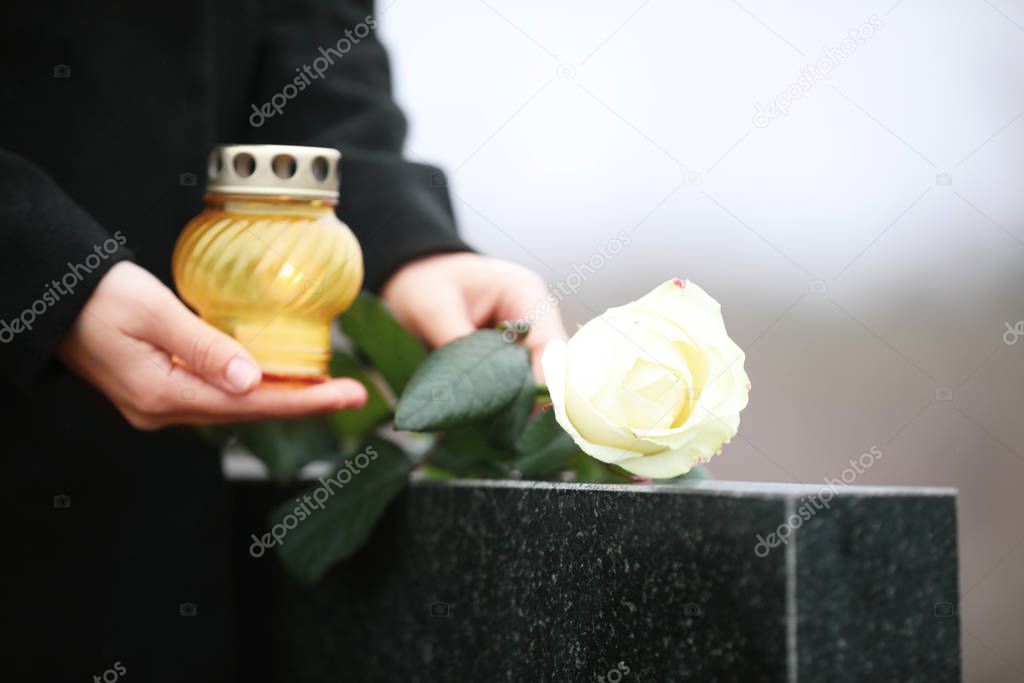 Woman with candle and rose near black granite tombstone outdoors