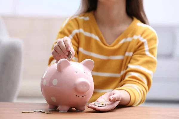 Woman putting coin into piggy bank at wooden table, closeup — Stock Photo, Image