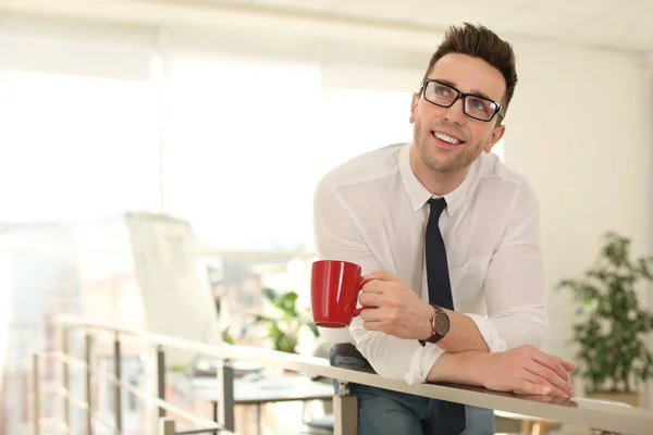 Jovem empresário com xícara de bebida relaxante no escritório durante o — Fotografia de Stock