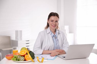 Nutritionist with clipboard and laptop at desk in office clipart