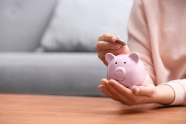 Woman putting coin into piggy bank at wooden table, closeup. Spa