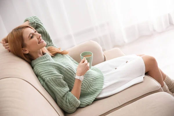 Giovane donna con tazza di bevanda rilassante sul divano di casa — Foto Stock