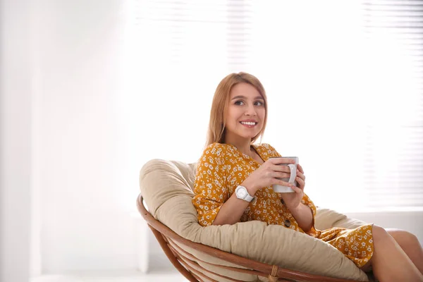 Mujer joven con taza de bebida relajante en silla de papasán cerca de ganar — Foto de Stock