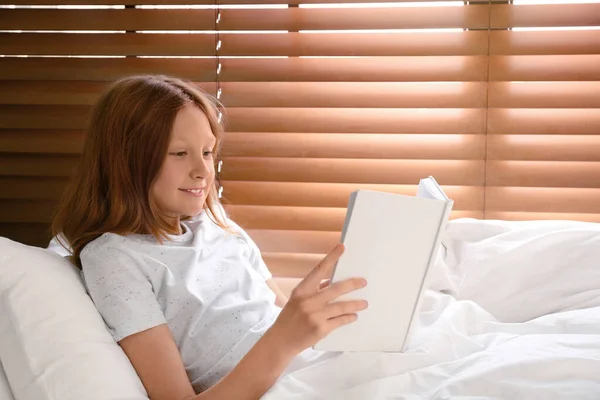 Cute preteen girl reading book in bed near window — Stock Photo, Image