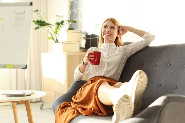 Young woman with cup of drink relaxing on couch at workplace
