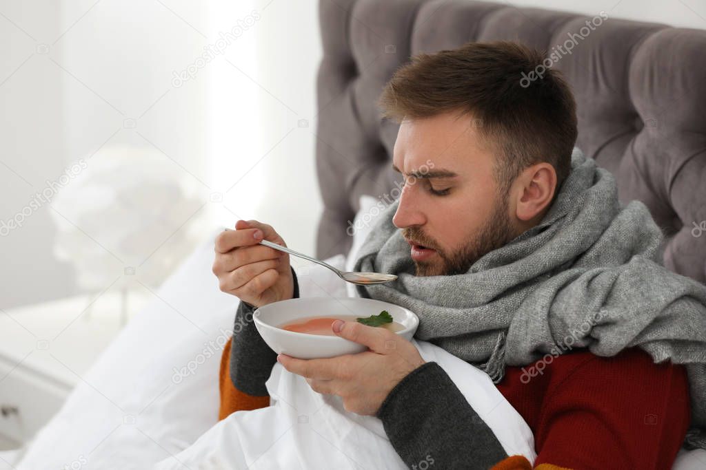 Sick young man with bowl of tasty soup in bed at home